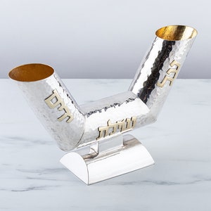 May include: A silver Kiddush cup with two spouts, each with a gold inscription. The cup is hammered and has a textured surface. The base is also silver and has a rounded shape.