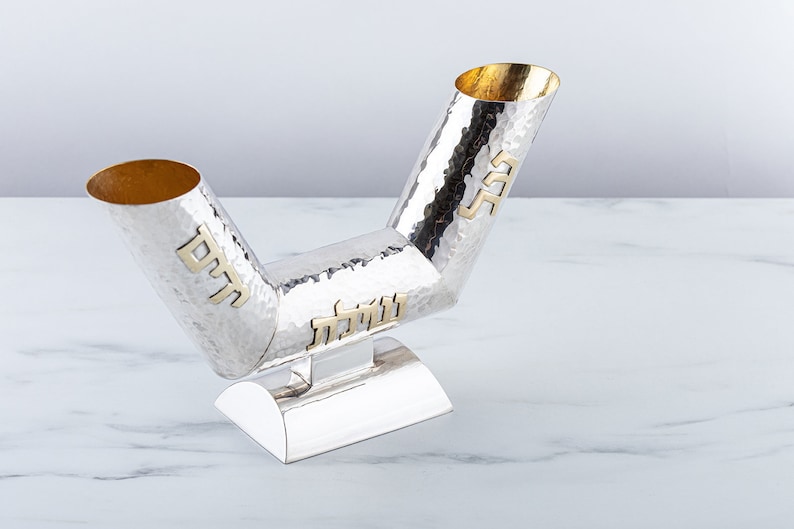 May include: A silver Kiddush cup with two spouts, each with a gold inscription. The cup is hammered and has a textured surface. The base is also silver and has a rounded shape.