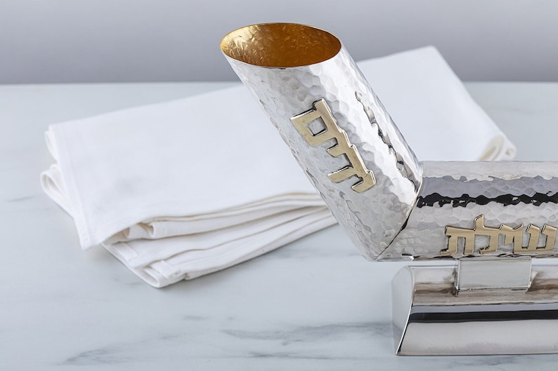 May include: A silver Kiddush cup with a hammered finish and gold Hebrew lettering. The cup is on a stand and is tilted to the side.