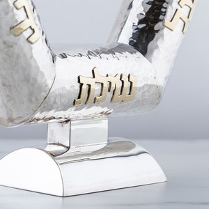 May include: A silver menorah with a hammered finish and gold Hebrew lettering. The menorah has a unique V-shaped design and a round base.