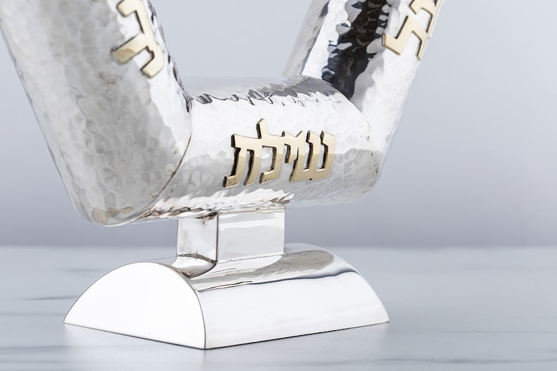 May include: A silver menorah with a hammered finish and gold Hebrew lettering. The menorah has a unique V-shaped design and a round base.