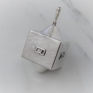 May include: A silver-colored dreidel featuring a hammered metal texture on its four sides. Each side displays a single raised Hebrew letter in a polished finish. The spinning top has a slender, tapered handle and a pointed base.