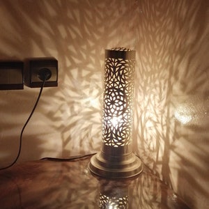 Pode incluir: Um candeeiro de metal dourado com um abajur cilíndrico que apresenta um design delicado e complexo de corte. O candeeiro projeta uma luz quente e com padrões na parede e na superfície da mesa.
