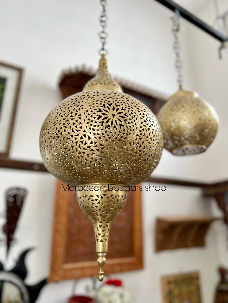 May include: Two gold Moroccan pendant lights with intricate cut-out designs. The lights are hanging from a black metal rod. The text "Moroccan bazaars shop" is visible in the image.
