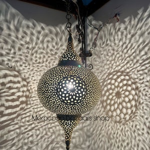 May include: A large, round, ornate, brass Moroccan pendant light with intricate cut-out designs. The light casts intricate shadows on the wall behind it.