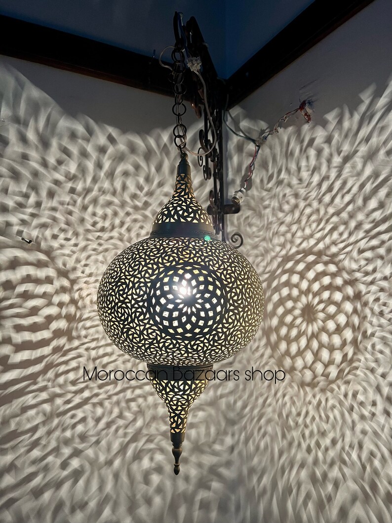 May include: A large, round, ornate, brass Moroccan pendant light with intricate cut-out designs. The light casts intricate shadows on the wall behind it.