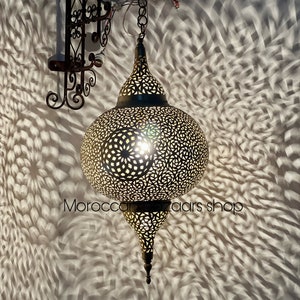 May include: A large, round, brass Moroccan lantern with intricate cut-out designs. The lantern is hanging from a chain and is lit from within, casting intricate shadows on the wall behind it. The text "Morocco Bazaar Shop" is visible in the lower right corner of the image.