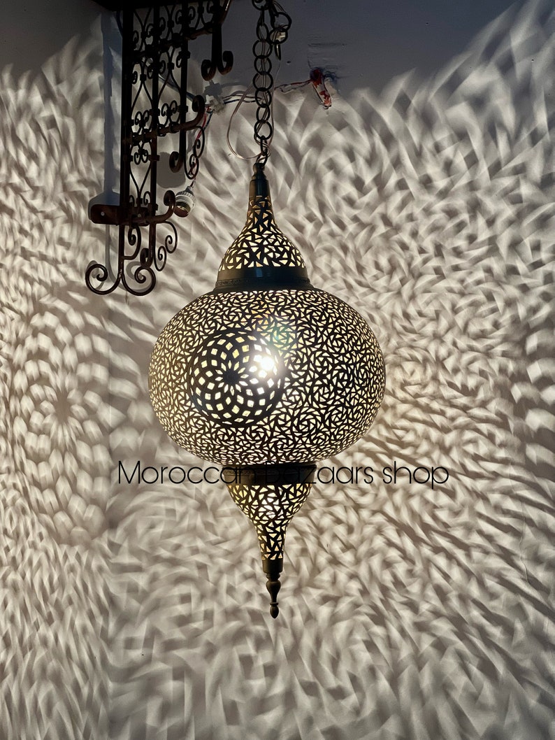 May include: A large, round, brass Moroccan lantern with intricate cut-out designs. The lantern is hanging from a chain and is lit from within, casting intricate shadows on the wall behind it. The text "Morocco Bazaar Shop" is visible in the lower right corner of the image.