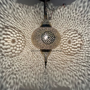 May include: A gold Moroccan pendant light with intricate cutouts casts a beautiful pattern of light and shadow on the wall. The light is hanging from a chain and has a pointed bottom.