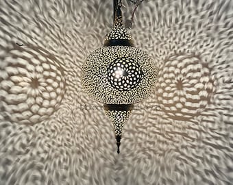 May include: A gold Moroccan pendant light with intricate cutouts casts a beautiful pattern of light and shadow on the wall. The light is hanging from a chain and has a pointed bottom.