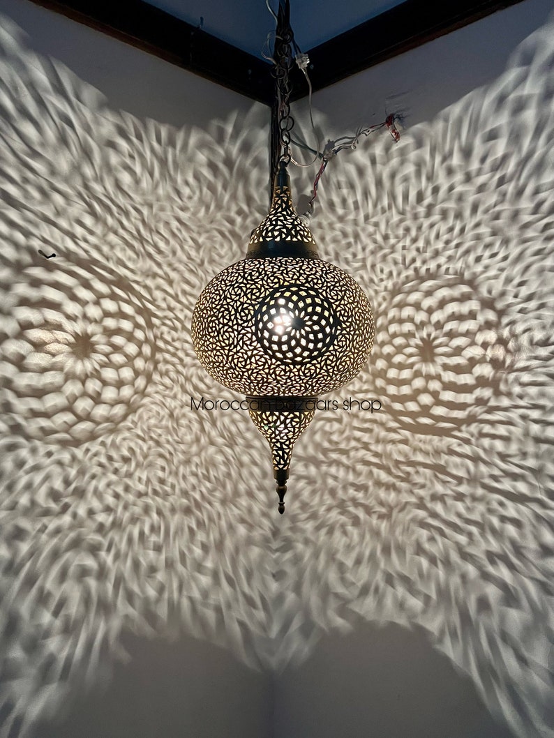 May include: A gold Moroccan pendant light with intricate cutouts casts a beautiful pattern of light and shadow on the wall. The light is hanging from a chain and has a pointed bottom.
