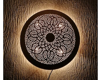 May include: A round, decorative wall lamp with a intricate, cut-out design. The lamp is made of metal and has a warm, golden finish. It features a circular pattern with a central circle and four smaller circles around it. The lamp is lit and casts a warm glow.
