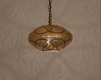 Może przedstawiać: Wisząca lampa z mosiądzu z okrągłym, perforowanym kloszem. Klosz ma ozdobny wzór z nakładających się na siebie kół i liści. Lampa jest zawieszona na łańcuchu.