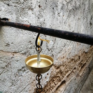 May include: A gold-coloured oil lamp hangs from a black metal bar attached to a rough, white wall. The lamp has a round bowl and a chain that connects it to the bar.