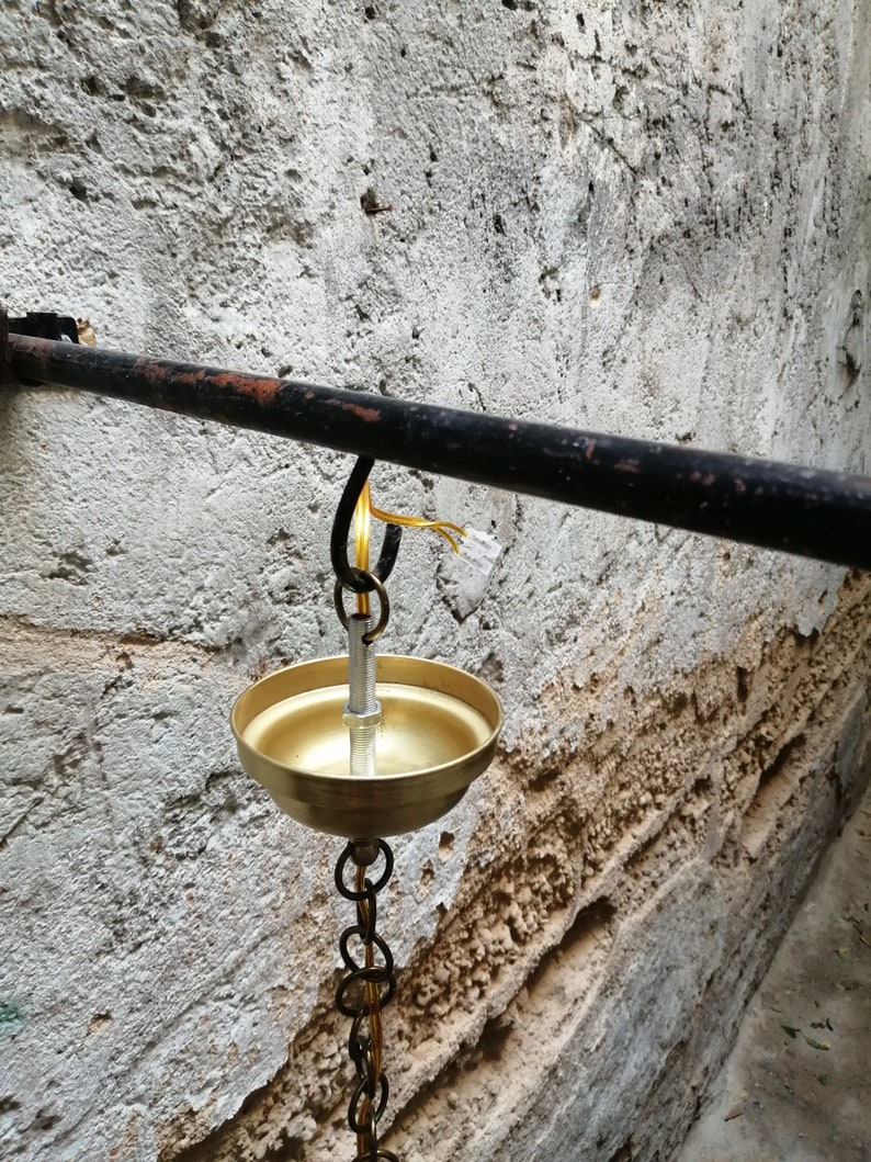 May include: A gold-coloured oil lamp hangs from a black metal bar attached to a rough, white wall. The lamp has a round bowl and a chain that connects it to the bar.