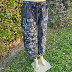 May include: Black and gray patterned pants with a drawstring waist. The pants feature a white mushroom design on the front and sides. The pants are displayed on a mannequin with a green grass background.
