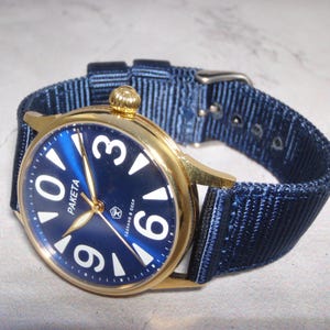 May include: A gold-toned wristwatch with a navy blue face and white numerals. The watch features a navy blue nylon band. The watch face has the word "PAKETA" printed on it. The watch is a vintage style.
