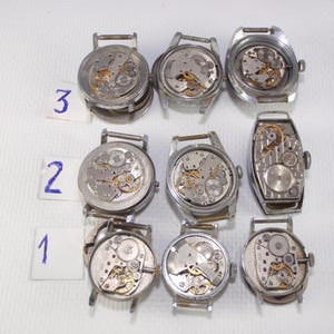 May include: A collection of disassembled vintage wristwatches, showcasing the intricate internal mechanisms. The watches are silver-toned, with visible gears, springs, and other components. Numbers 1, 2, and 3 are written on small white cards. The watches are displayed on a white surface.