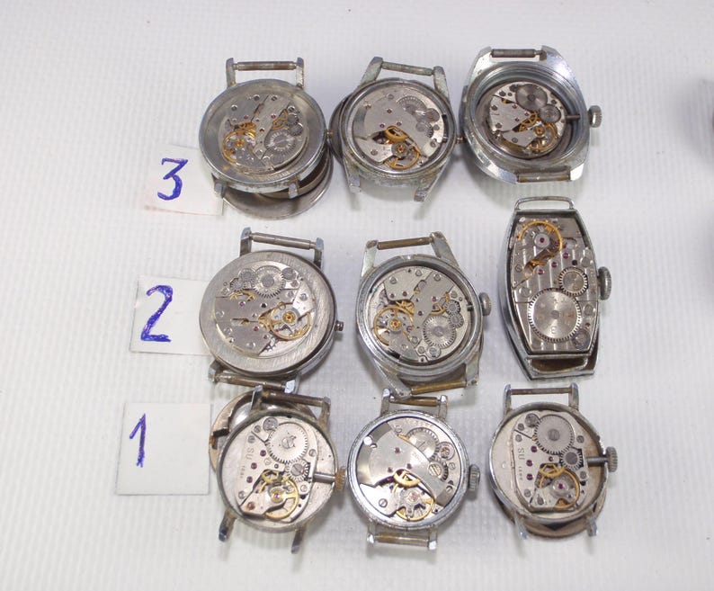 May include: A collection of disassembled vintage wristwatches, showcasing the intricate internal mechanisms. The watches are silver-toned, with visible gears, springs, and other components. Numbers 1, 2, and 3 are written on small white cards. The watches are displayed on a white surface.