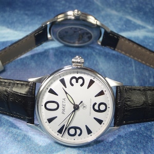 May include: A silver-tone wristwatch with a white dial, black Arabic numerals at 0, 3, 6, and 9, and a black textured leather strap. The dial features the text PAKETA and СДЕЛАНО В СССР. A second watch is visible in the background, showing its case back.