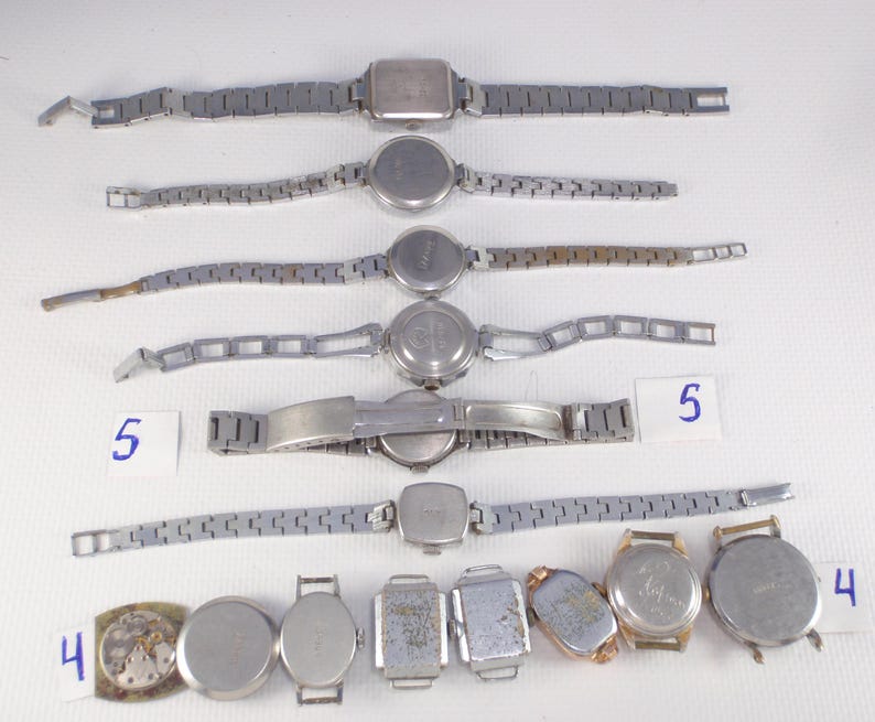 May include: A collection of vintage wristwatches with silver-toned metal bands and watch faces. The watches have various shapes, including round, square, and rectangular. Some watches are complete, while others are missing parts. Numbers 4 and 5 are written on small white cards.