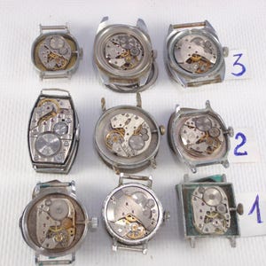 May include: A collection of nine vintage watch movements, showcasing intricate mechanical components. The watch cases are silver-toned and in various shapes, including round, square, and rectangular. The internal gears and springs are visible, with some labeled with numbers.