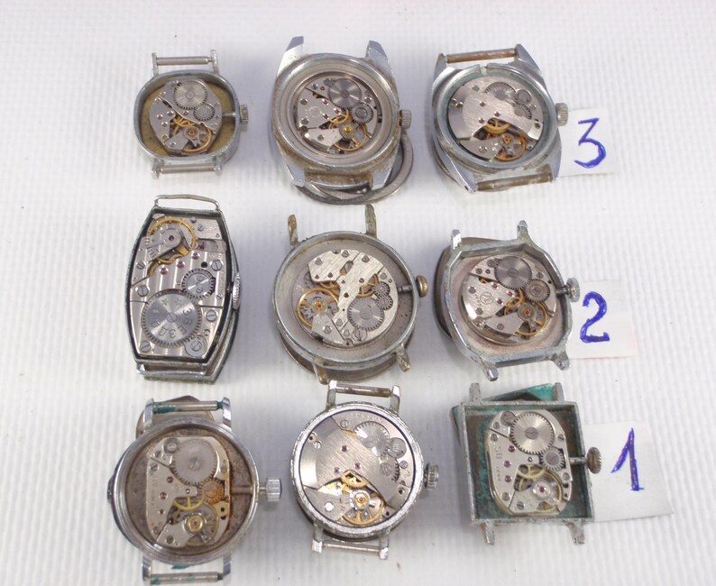 May include: A collection of nine vintage watch movements, showcasing intricate mechanical components. The watch cases are silver-toned and in various shapes, including round, square, and rectangular. The internal gears and springs are visible, with some labeled with numbers.