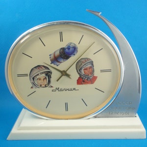Könnte beinhalten: Eine silberne und weiße Tischuhr mit einem runden Zifferblatt mit einem Weltraum-Design. Das Zifferblatt der Uhr zeigt ein Bild eines Raumschiffs, zwei Astronauten und den Text "Маяииа". Der Sockel der Uhr trägt den Text "CCCP BOCTON-1 12.IV.1961".