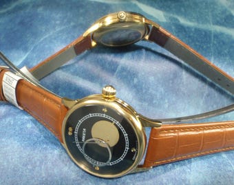 Op de afbeelding: Twee goudkleurige polshorloges met bruine leren bandjes worden getoond. Eén horloge heeft een zwarte wijzerplaat met een maanfase-ontwerp en het woord "RAKETA". Het andere horloge is ondersteboven en toont de achterkant. De horloges liggen op een blauw oppervlak.