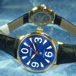 May include: Two gold-toned wristwatches with black leather bands are displayed. The watch in the foreground has a blue face with white numerals and the word "PAKETA". The second watch is in the background, showing the watch mechanism.