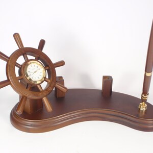 May include: A wooden desk organizer with a ship's wheel clock, a pen holder, and a pen stand. The organizer is made of dark brown wood and has a curved base.
