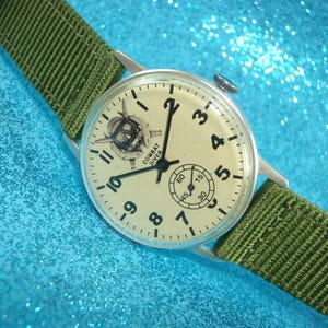 Puede incluir: Un reloj de pulsera plateado con una esfera blanca y agujas negras. El reloj tiene una correa de nylon verde y dice "Combat Diver" en la esfera.