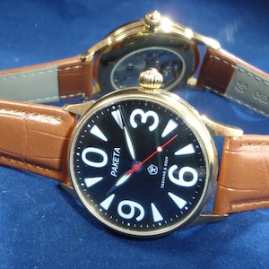 May include: Two gold wristwatches with brown leather bands. The watch on the left has a black face with white Arabic numerals and a red second hand. The watch on the right has a clear face with a visible mechanical movement.