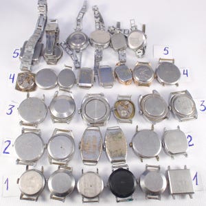 May include: A collection of vintage wristwatches with silver-toned metal bands and watch faces. The watches vary in shape, including round, oval, and rectangular designs. Some watches show signs of wear and tear, with visible internal mechanisms on a few.