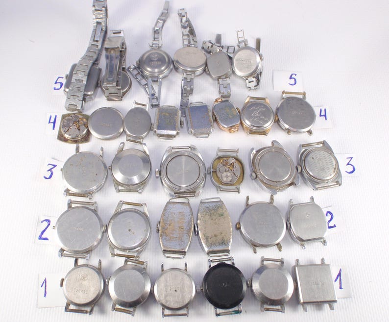 May include: A collection of vintage wristwatches with silver-toned metal bands and watch faces. The watches vary in shape, including round, oval, and rectangular designs. Some watches show signs of wear and tear, with visible internal mechanisms on a few.
