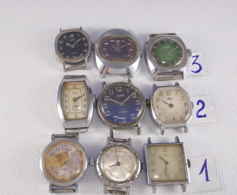 May include: Nine vintage wristwatches with various face designs and metal bands are displayed. The watch faces feature different colors, including black, green, and cream. Some watches have Arabic numerals, while others have a minimalist design.