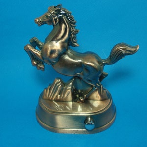 May include: A bronze-coloured metal horse figurine standing on a rocky base. The horse is rearing up on its hind legs with its front legs raised. The base has a round, decorative platform.