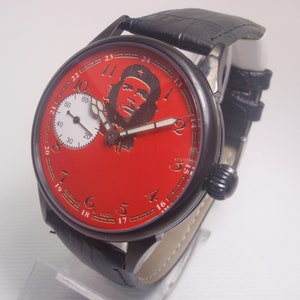 以下が含まれることがあります： 赤い文字盤と白い小文字盤を持つ黒い腕時計。文字盤には、赤い星がついたベレー帽をかぶったチェ・ゲバラの肖像画が描かれている。文字盤の縁には、1から12までの数字が金色で書かれている。時計は黒い革のバンドが付いている。