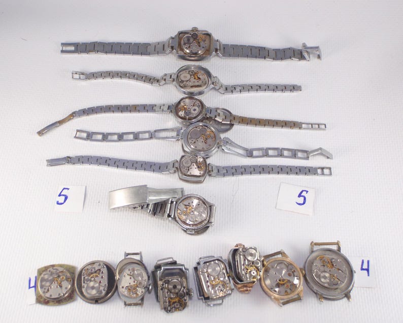 May include: A collection of vintage wristwatches, some with exposed mechanical movements. The watches have silver-toned metal bands and various case shapes, including square and rectangular. Some watches are missing parts. Numbers 4 and 5 are written on small white cards.