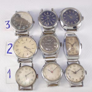 May include: Nine vintage wristwatches with silver-tone metal bands and various face designs. The watch faces include white, blue, and brown tones, with visible numbers and hands. Some watches have the brand name "Zaria" on the face.