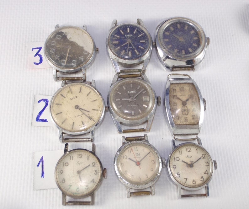 May include: Nine vintage wristwatches with silver-tone metal bands and various face designs. The watch faces include white, blue, and brown tones, with visible numbers and hands. Some watches have the brand name "Zaria" on the face.