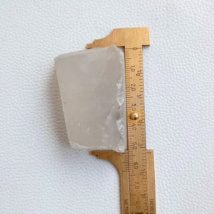 May include: A translucent, irregularly shaped crystal is measured with a brass ruler. The ruler shows measurements in inches. The crystal appears to be approximately 3 inches in length.
