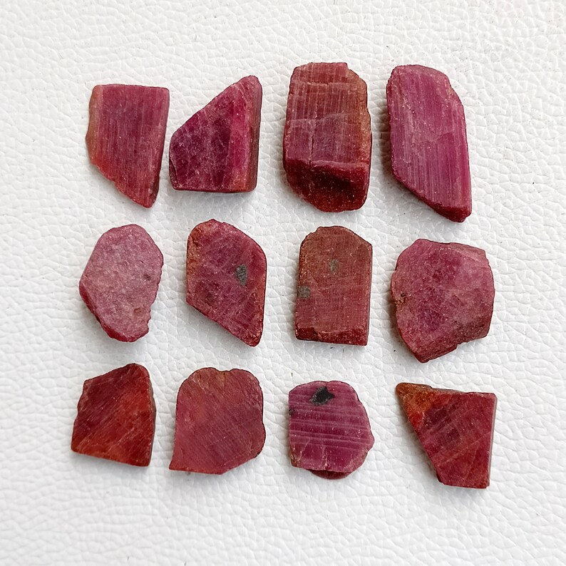 May include: A collection of twelve rough, unpolished ruby gemstones. The rubies are various shades of red and pink, and have a natural, irregular shape.