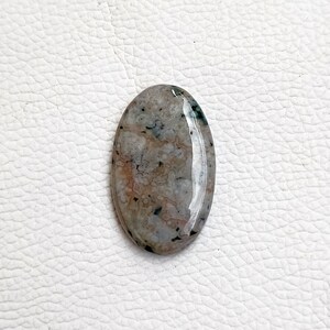 May include: A smooth, oval-shaped, light gray agate cabochon with brown and green inclusions. The stone has a polished surface and is translucent.