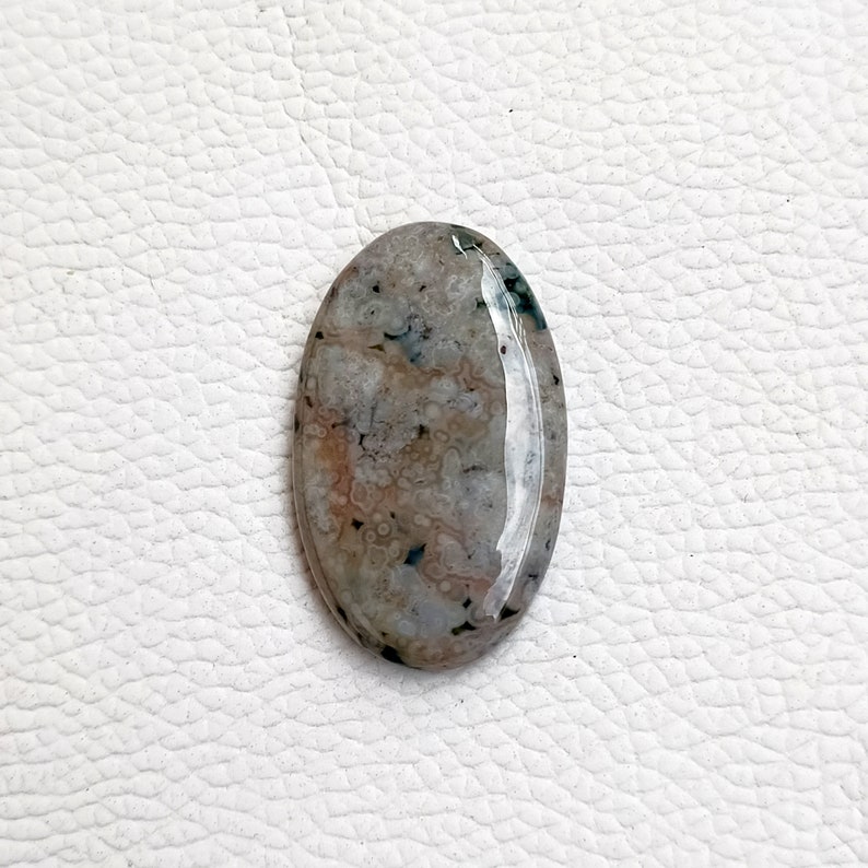 May include: A smooth, oval-shaped, light gray agate cabochon with brown and green inclusions. The stone has a polished surface and is translucent.