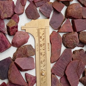 May include: A collection of rough, red ruby gemstones of various sizes and shapes, measured with a ruler in inches. The ruler is made of wood and has markings for both inches and centimetres.