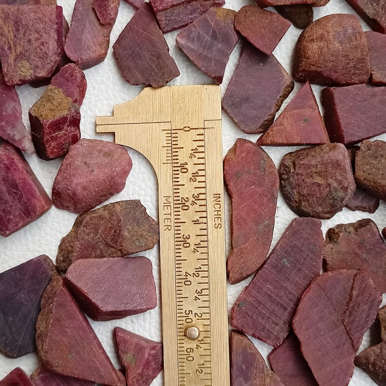 May include: A collection of rough, red ruby gemstones of various sizes and shapes, measured with a ruler in inches. The ruler is made of wood and has markings for both inches and centimetres.