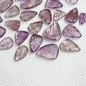 May include: A collection of purple and clear amethyst gemstone teardrop shapes with carved details. The stones are arranged on a white textured surface.