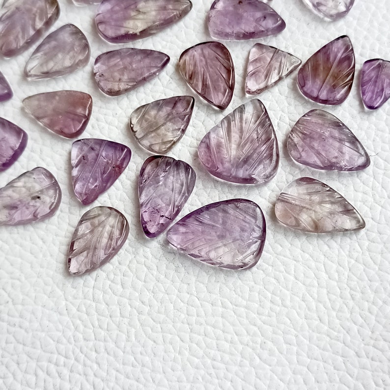 May include: A collection of purple and clear amethyst gemstone teardrop shapes with carved details. The stones are arranged on a white textured surface.