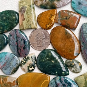 May include: A collection of polished, natural, green, brown, and red agate stones. The stones are various shapes and sizes, including oval, teardrop, and rectangular.  The stones are arranged on a white surface.  One stone is next to a US quarter dollar coin.
